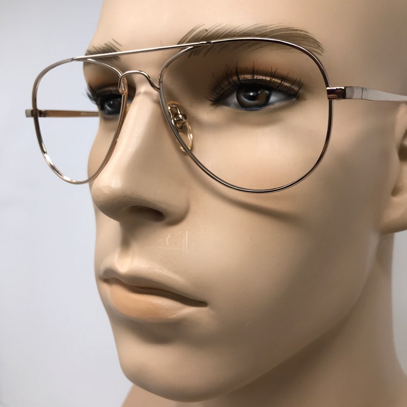 May include: Gold-tone aviator eyeglasses with clear lenses. The frames feature a double bridge and a classic teardrop shape. The glasses are displayed on a mannequin.