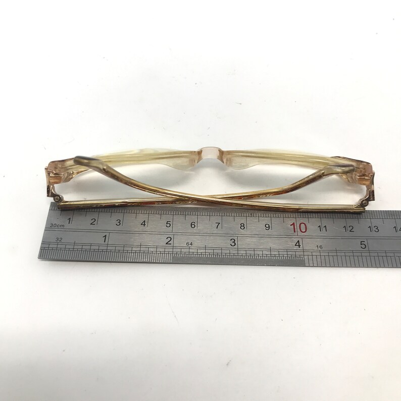 May include: A pair of eyeglasses with a light brown frame and clear lenses. The glasses are measured with a ruler, showing a length of approximately 5 inches. The frame has a classic design.