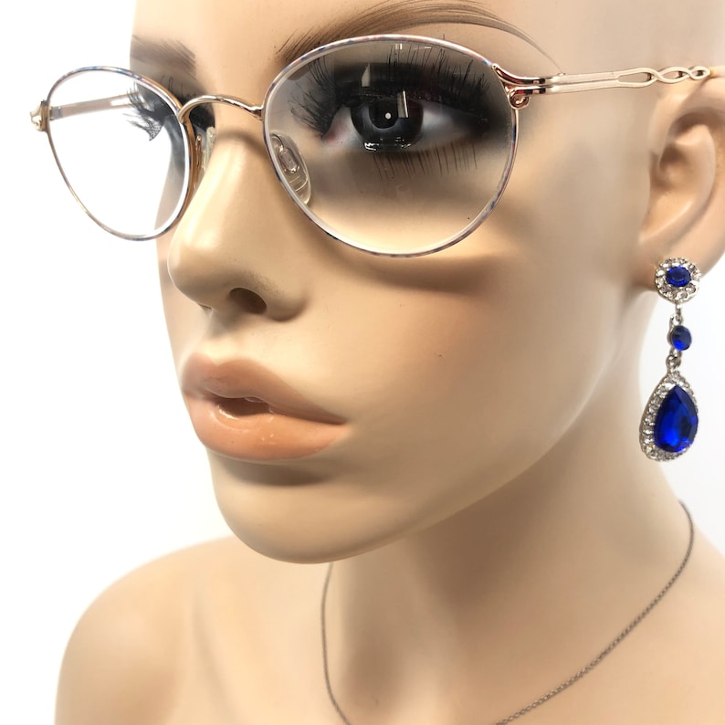 May include: Close-up of a mannequin wearing gold-rimmed eyeglasses and blue drop earrings. The eyeglasses have a thin gold frame and clear lenses. The earrings feature a large blue teardrop-shaped stone surrounded by smaller clear stones.