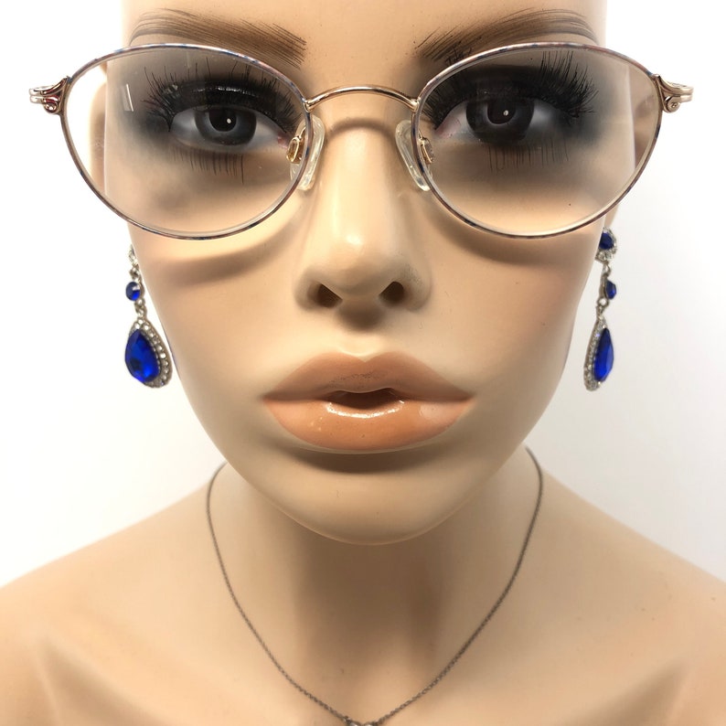 May include: Gold-rimmed eyeglasses with oval lenses are worn by a mannequin. The mannequin also wears dangling blue teardrop earrings with silver accents and a delicate silver necklace. The background is white.