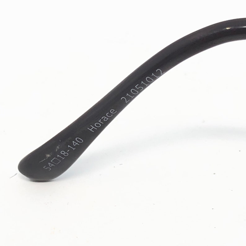 May include: Close-up of the temple of a pair of black eyeglasses. The text "54018-140 Horace 21051012" is printed on the temple arm. The arm is curved and has a matte finish.