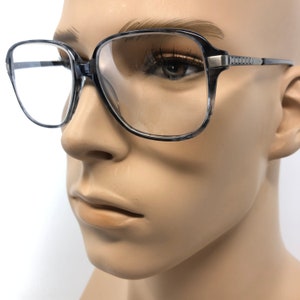 May include: Square-framed spectacles with a marbled grey and black pattern. The glasses have a silver-toned metal accent on the temples and a clear lens. The spectacles are displayed on a mannequin head.