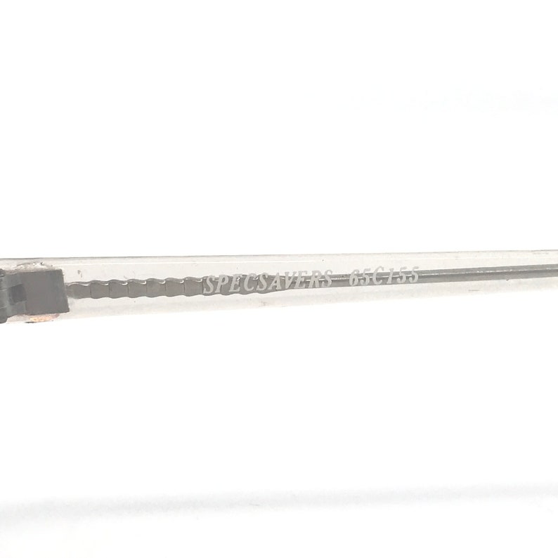 May include: Close-up of a clear eyeglass frame arm with the text "SPECSAVERS 65 C155" printed in a light gray color. The frame arm is made of a transparent material and has a rectangular shape.