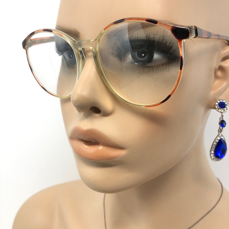 K&ouml;nnte beinhalten: Brille mit transparentem Rahmen und Schildpattmuster an den oberen R&auml;ndern und B&uuml;geln. Die Brille ist rund und &uuml;bergro&szlig;. Ein Schaufensterpuppenkopf tr&auml;gt die Brille und ein Paar blaue Tr&auml;nenohrringe.