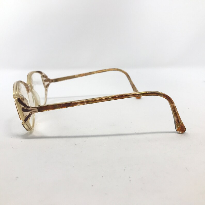 May include: Vintage eyeglasses with a clear, octagonal frame and gold-toned metal arms. The arms have a slight curve and are attached to the frame with a decorative element. The lenses are clear, and the glasses are set against a white background.