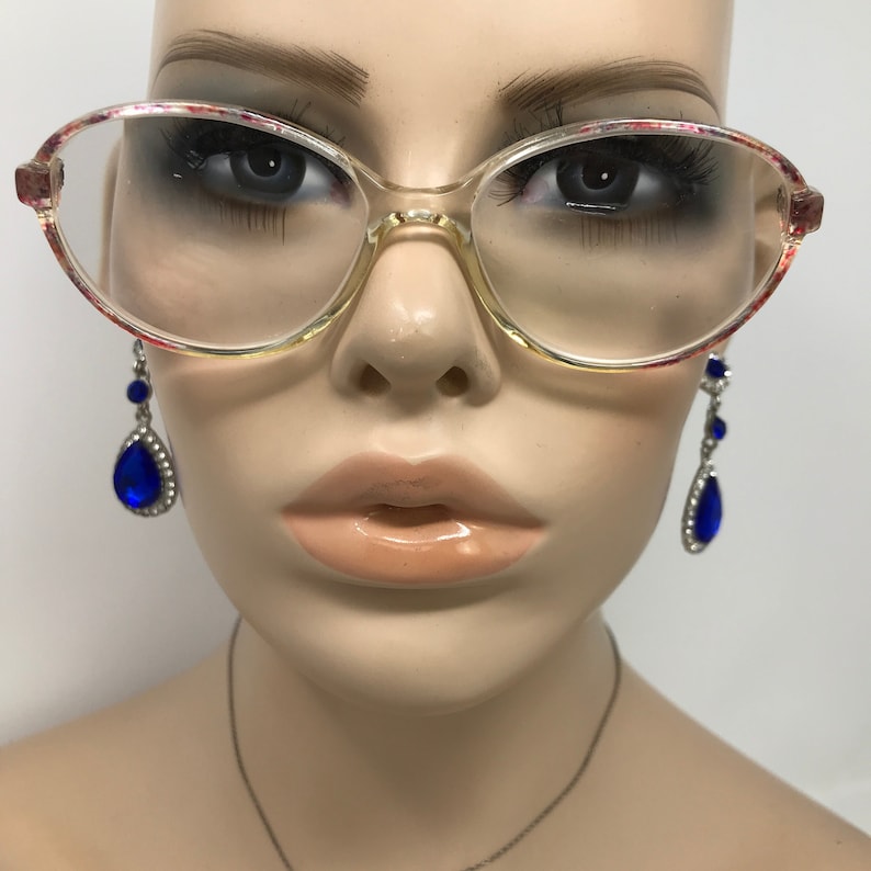 May include: Clear oval eyeglasses with a floral pattern on the upper rims. The glasses are set on a mannequin head, with blue teardrop earrings hanging on either side.
