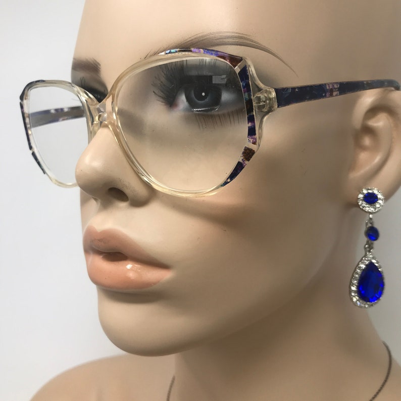 Puede incluir: Gafas con montura transparente, patillas estampadas y forma cuadrada. Las gafas est&aacute;n sobre una cabeza de maniqu&iacute;, con un par de pendientes colgantes de piedras preciosas azules. Las gafas tienen una est&eacute;tica retro y vintage.