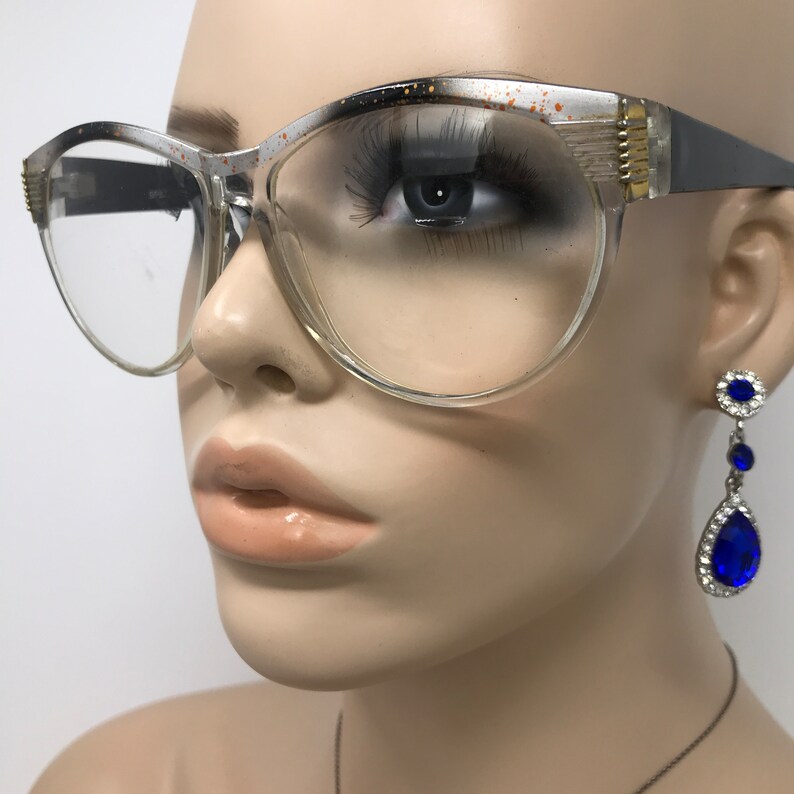 May include: Clear-framed eyeglasses with a gray and gold accent on the upper frame. The glasses have a cat-eye shape. A mannequin wears the glasses and a pair of blue teardrop earrings.