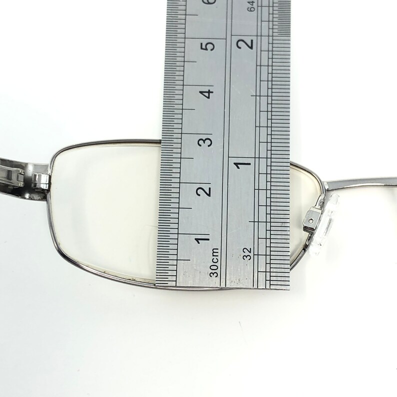 May include: Silver-framed eyeglasses with clear lenses are measured with a ruler. The ruler shows the glasses are approximately 3 inches wide. The glasses have a rectangular shape and a hinge design.