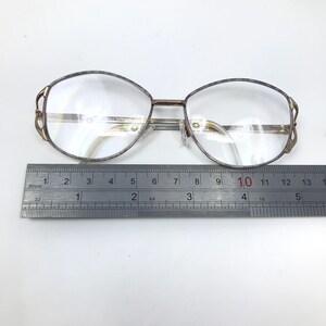 Op de afbeelding: Vintage bril met een goudkleurig metalen montuur en heldere glazen. De ovale monturen hebben een decoratief ontwerp op de veren. Een liniaal eronder toont een meting van ongeveer 13 cm.