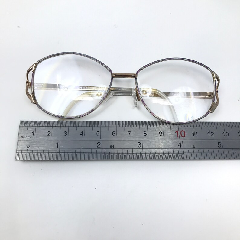 Op de afbeelding: Vintage bril met een goudkleurig metalen montuur en heldere glazen. De ovale monturen hebben een decoratief ontwerp op de veren. Een liniaal eronder toont een meting van ongeveer 13 cm.