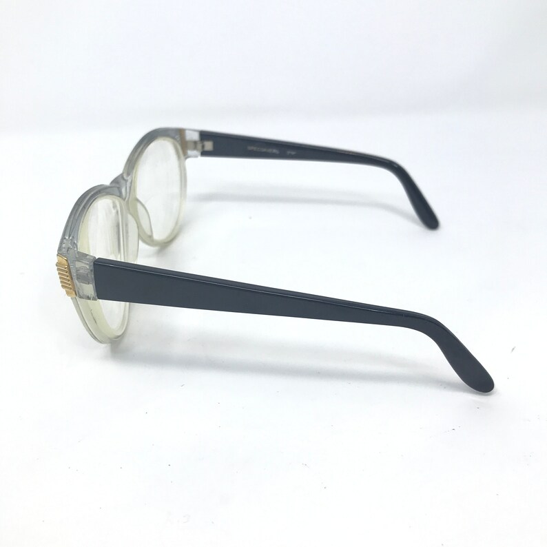 May include: Clear plastic framed glasses with black arms. The glasses have a rounded shape and gold accents on the frame.