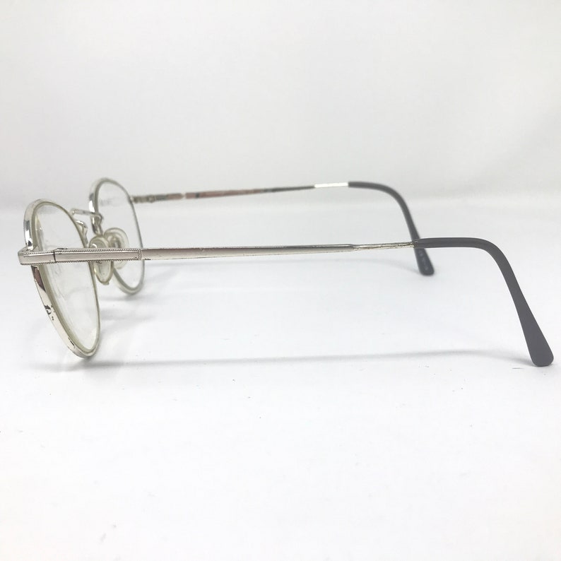 Peut inclure: Lunettes &agrave; monture argent&eacute;e avec verres ronds. Les lunettes ont une monture fine argent&eacute;e avec un pont et des plaquettes nasales. Les branches sont argent&eacute;es et passent au noir aux extr&eacute;mit&eacute;s. Les verres sont clairs.