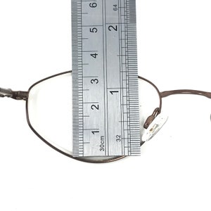 Puede incluir: Gafas con montura met&aacute;lica marr&oacute;n con una regla colocada sobre la lente. La regla muestra una medida de aproximadamente 5 cm. Las gafas tienen un dise&ntilde;o cl&aacute;sico y minimalista con lentes transparentes y almohadillas nasales.