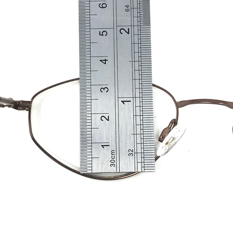 Puede incluir: Gafas con montura met&aacute;lica marr&oacute;n con una regla colocada sobre la lente. La regla muestra una medida de aproximadamente 5 cm. Las gafas tienen un dise&ntilde;o cl&aacute;sico y minimalista con lentes transparentes y almohadillas nasales.