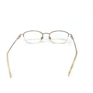 May include: A pair of eyeglasses with a gold-colored metal frame. The lenses are clear, and the arms extend outward with a curved design. The glasses are set against a white background.
