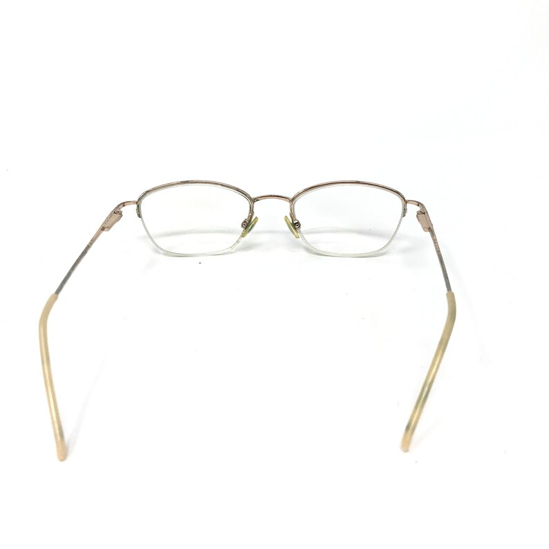 May include: A pair of eyeglasses with a gold-colored metal frame. The lenses are clear, and the arms extend outward with a curved design. The glasses are set against a white background.