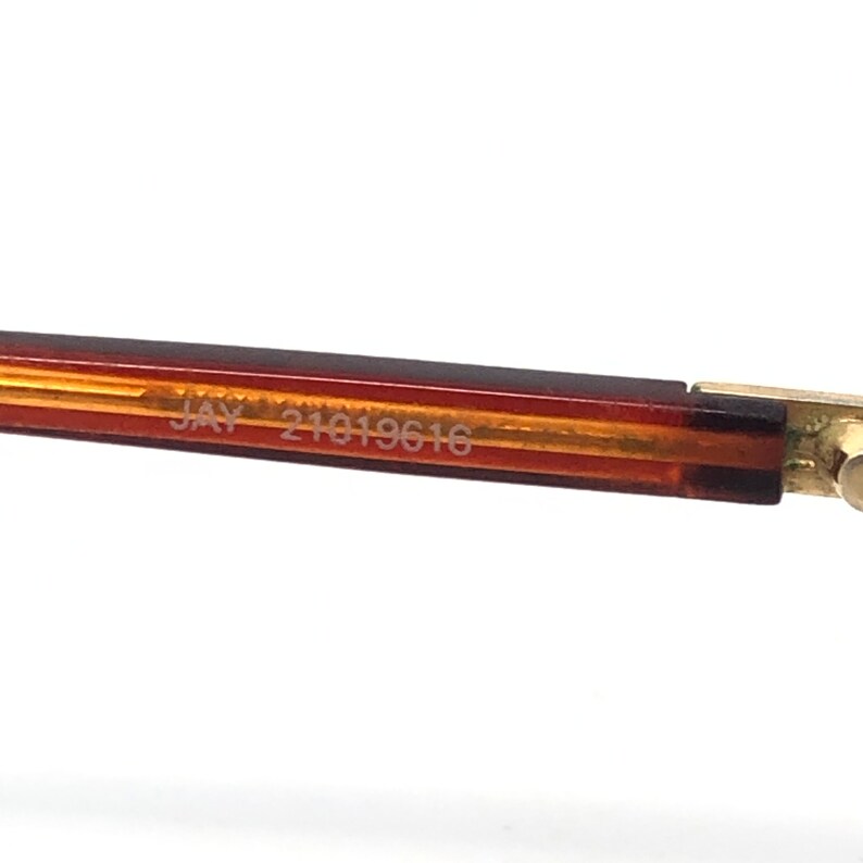 May include: Close-up of the arm of a pair of eyeglasses. The frame is a gradient of brown and gold with the text "JAY 21019616" printed on the arm. The end of the arm has a gold-colored hinge.