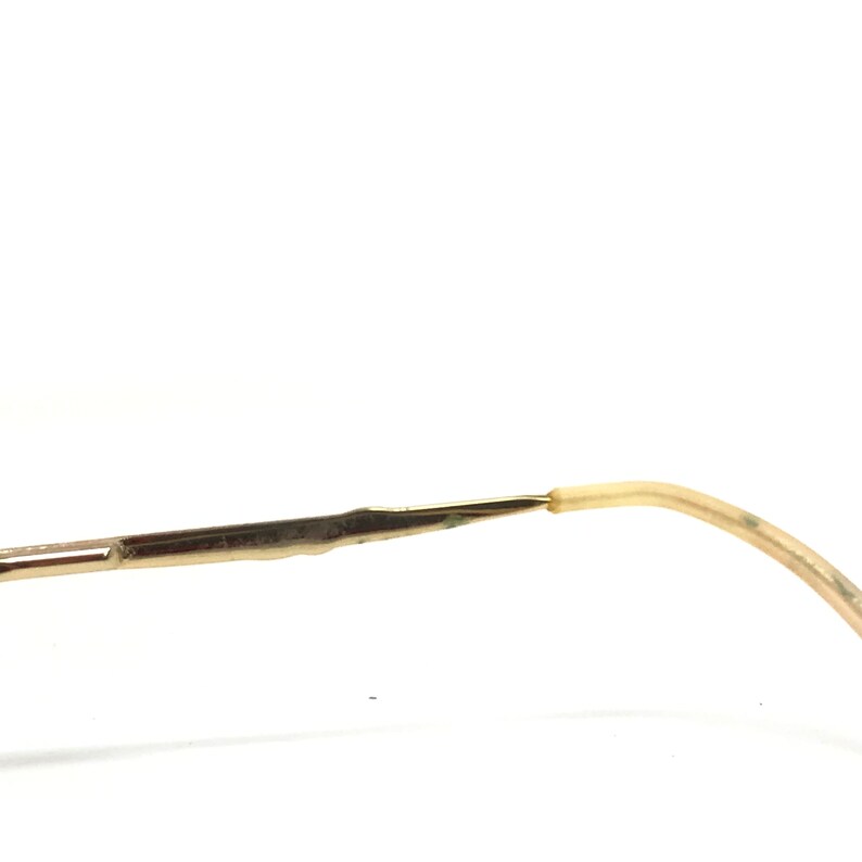 May include: Close-up of a pair of eyeglasses. The frame is a light gold color, with a thin, delicate design. The earpiece is a light beige color. The glasses are set against a white background.