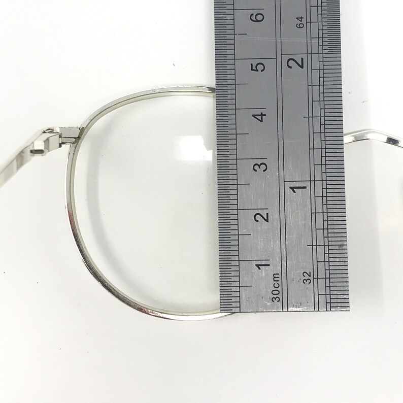 Peut inclure: Gros plan sur une paire de lunettes &agrave; monture argent&eacute;e avec des verres transparents. Une r&egrave;gle est plac&eacute;e &agrave; c&ocirc;t&eacute; des lunettes, indiquant une mesure d'environ 5 cm. Les lunettes ont une forme ronde classique.
