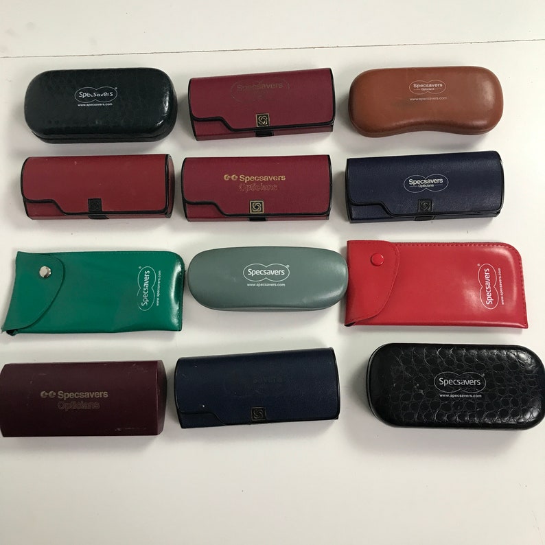 May include: A collection of various colored Specsavers eyeglass cases. The cases are in black, red, brown, blue, green, and gray. Each case has the Specsavers logo printed on it. The cases are made of leather or a leather-like material.