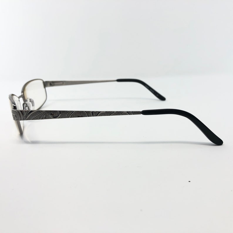 May include: Silver-toned eyeglasses with a decorative floral pattern on the temples. The frames have a rectangular shape with clear lenses. The earpieces are black and curved for a comfortable fit. The glasses are set against a white background.