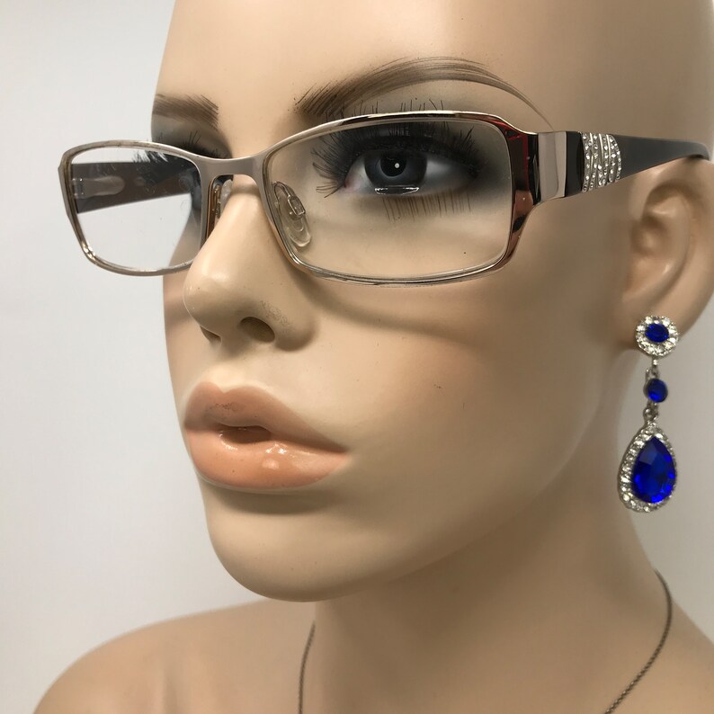 May include: Eyeglasses with a silver frame and rectangular lenses. The temples are black with a silver accent and rhinestones. A mannequin head is wearing the glasses and a pair of blue and silver drop earrings.