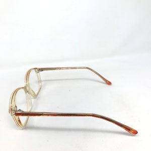May include: Clear eyeglasses with a light brown and red accent on the frame. The glasses have a rectangular shape and thin temples. The lenses are clear, and the overall design is vintage.