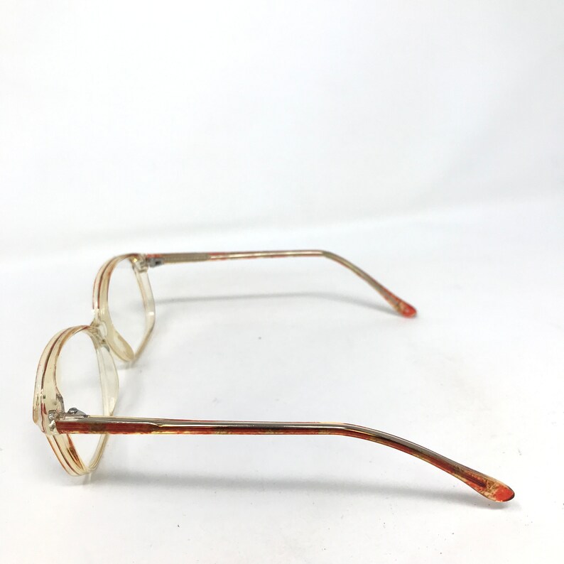 May include: Clear eyeglasses with a light brown and red accent on the frame. The glasses have a rectangular shape and thin temples. The lenses are clear, and the overall design is vintage.