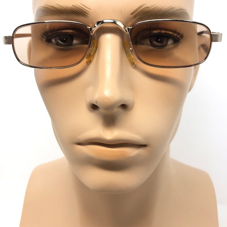 May include: Rectangular sunglasses with a gold-colored metal frame and amber-tinted lenses. The glasses have a classic design, with a bridge and temples. The sunglasses are displayed on a mannequin head.