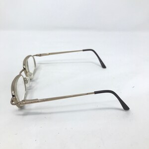 May include: Silver-toned eyeglasses with oval lenses and thin temples. The earpieces are black. The glasses are set against a white background. The frames have a classic, minimalist design.