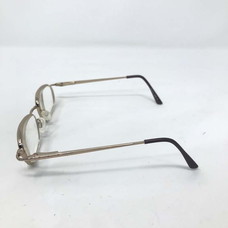 May include: Silver-toned eyeglasses with oval lenses and thin temples. The earpieces are black. The glasses are set against a white background. The frames have a classic, minimalist design.