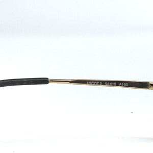 May include: Close-up of the arm of a pair of eyeglasses. The frame is gold-colored with the text "ASCOT 2 54-15 A160" printed on the arm. The earpiece is black.