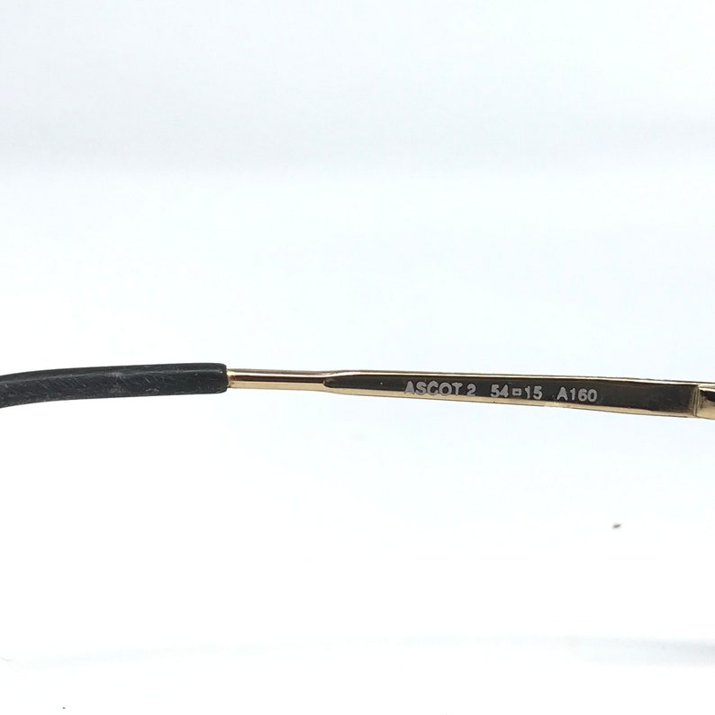 May include: Close-up of the arm of a pair of eyeglasses. The frame is gold-colored with the text "ASCOT 2 54-15 A160" printed on the arm. The earpiece is black.