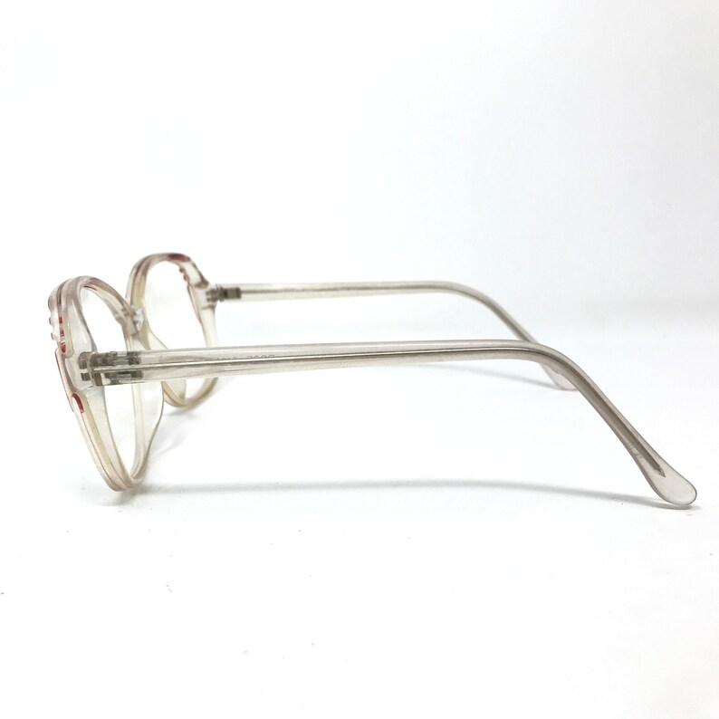 May include: Clear, vintage eyeglasses with a light brown tint. The frames are a light, translucent color with a rounded shape. The temples are thin and extend to the ear pieces. The glasses are set against a white background.