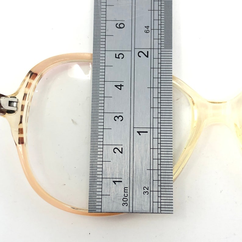 Puede incluir: Gafas vintage con montura marr&oacute;n claro y lentes transparentes. La montura se mide con una regla, mostrando un ancho de aproximadamente 13 cm. Las gafas tienen un dise&ntilde;o cl&aacute;sico y retro.