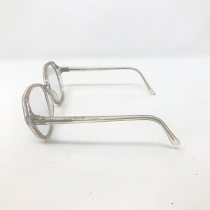 May include: Two pairs of clear, octagonal eyeglasses with thin, transparent frames. The glasses are positioned on a white surface, showcasing their side profile. The lenses are clear, and the temples curve gently.