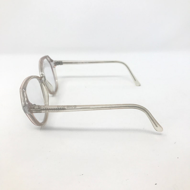 May include: Two pairs of clear, octagonal eyeglasses with thin, transparent frames. The glasses are positioned on a white surface, showcasing their side profile. The lenses are clear, and the temples curve gently.