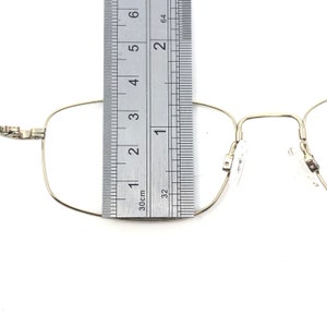 May include: Close-up of a pair of gold-colored eyeglasses with a ruler placed over the frame. The ruler shows measurements in inches. The glasses have a rectangular shape and clear lenses. The frame is thin and delicate.