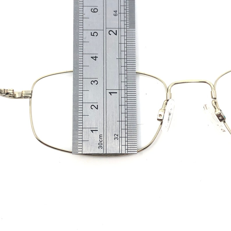 May include: Close-up of a pair of gold-colored eyeglasses with a ruler placed over the frame. The ruler shows measurements in inches. The glasses have a rectangular shape and clear lenses. The frame is thin and delicate.