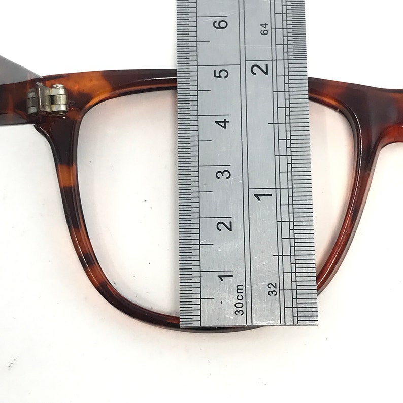 May include: Close-up of eyeglasses with a tortoise shell pattern. A ruler is placed over the frame, showing measurements. The glasses have a rectangular shape and a hinge detail. The frame is approximately 1 inch wide.