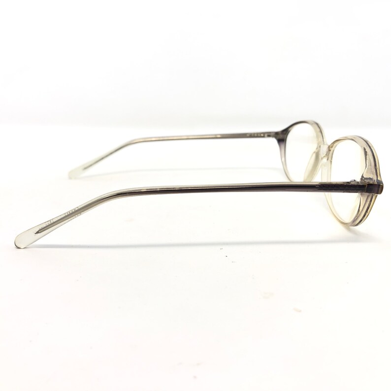May include: Clear, oval-shaped eyeglasses with a translucent frame and thin, silver-toned temples. The glasses are viewed from the side, showcasing the lens shape and the delicate design of the arms. The overall aesthetic is minimalist and modern.