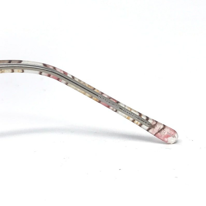 May include: Close-up of a patterned eyeglass arm. The arm features a floral design in pink, white, and tan. The text "Sassavare 100046" is printed on the arm. The end of the arm is clear.