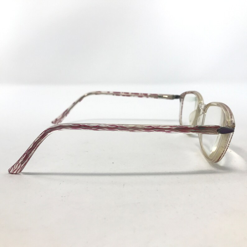 May include: A pair of eyeglasses with clear lenses and a light-colored frame. The temples feature a textured pattern with red and white stripes. The frame has a subtle, translucent appearance, and the lenses are clear.