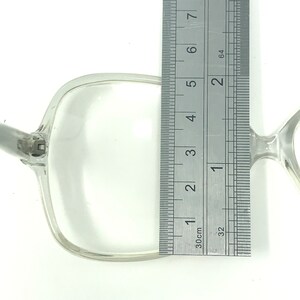 May include: Clear, square-framed eyeglasses with a silver hinge. The frame measures approximately 2 inches in height, as indicated by the ruler in the image. The glasses are set against a white background.