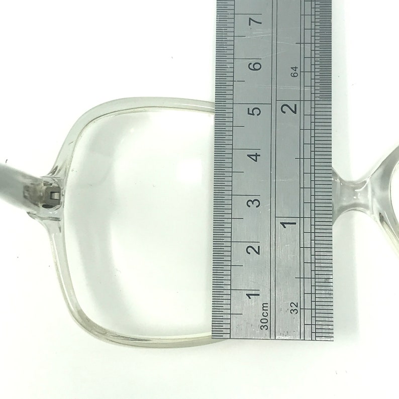 May include: Clear, square-framed eyeglasses with a silver hinge. The frame measures approximately 2 inches in height, as indicated by the ruler in the image. The glasses are set against a white background.