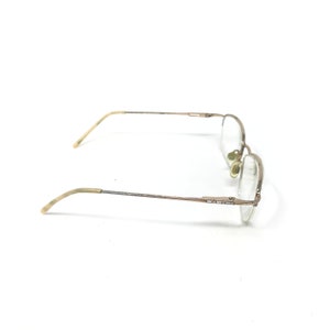 May include: A pair of eyeglasses with a thin, gold-colored metal frame. The lenses are clear, and the temples have a light-colored, curved design. The glasses are set against a white background.