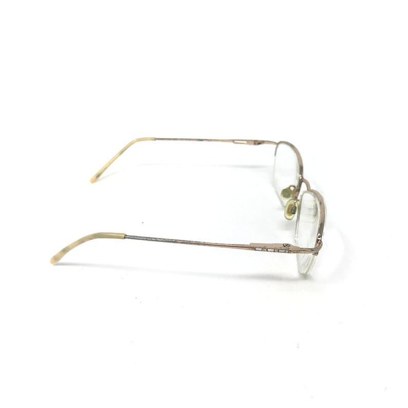 May include: A pair of eyeglasses with a thin, gold-colored metal frame. The lenses are clear, and the temples have a light-colored, curved design. The glasses are set against a white background.