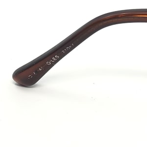 May include: Close-up of the arm of a pair of brown eyeglasses. The text "GILES" is printed on the arm, along with other numbers. The arm is a solid, dark brown color.