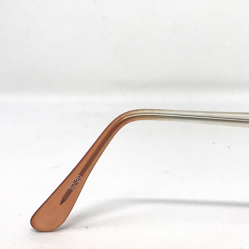 May include: Close-up of eyeglasses arm with a light brown, curved temple and a silver-colored arm. The text "52-20" is printed on the temple. The glasses are set against a white background.